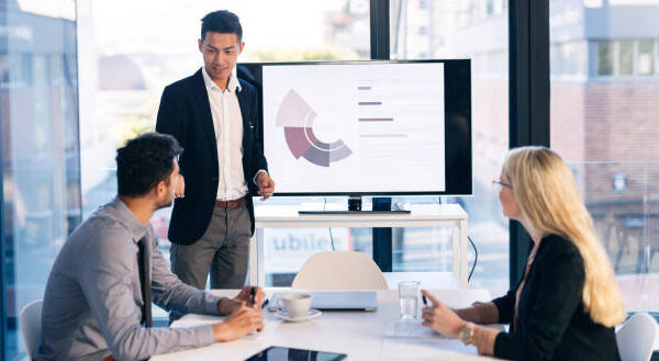A group of motivated young professionals engage in a dynamic business meeting in a modern office setting. A presenter shares data insights on a screen, fostering collaboration and strategic discussions. The office features large windows, providing a bright and open atmosphere that enhances creativity and communication.