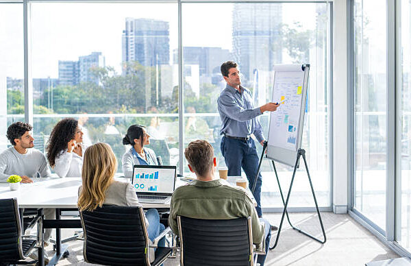 Business people watching a presentation on the whiteboard. A man is writing on the whiteboard with charts and graphs. They are sitting in a board room, there are laptop computers and technology on the table. Finance charts and graphs can be seen. There is a window behind him with city views.