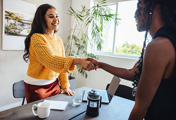 Two professional women in their thirties, shaking hands as part of a meeting in an architecture studio environment, symbolizing collaboration, respect, and teamwork in a professional setting.