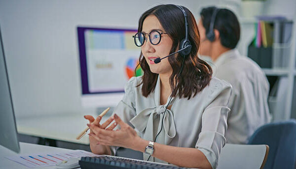 A professional Asian female call center agent wearing a headset works at a computer workstation while reviewing documents, captured with a partial POV perspective including a blurred colleague in the foreground. The scene represents modern customer support operations, remote communication, teamwork, and online service assistance in a corporate office environment.