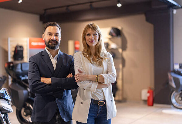 Two professional adults stand with arms crossed in a stylish showroom, motorcycles visible in the background. The scene conveys teamwork, confidence, and a polished business environment.
