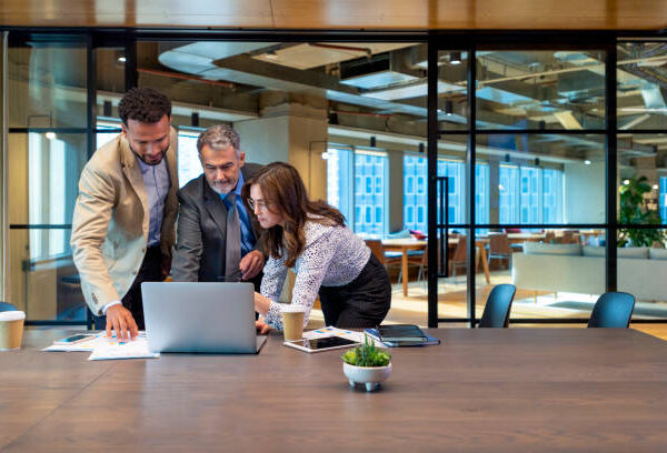 Business team working on a laptop computer. Three people are wearing business clothing. They are standing in a board room. Multi ethnic group with Caucasian and black men and women. There is other technology and paperwork on the conference table
